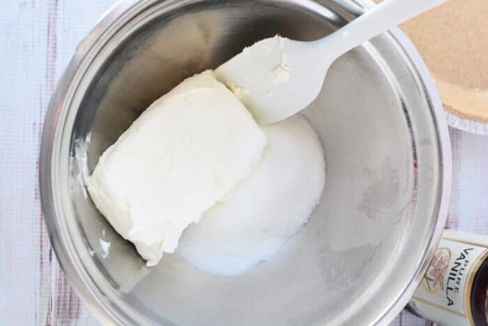 A rubber spatula mixing a block of cream cheese and white sugar for a 3 ingredient cheesecake in a bowl.