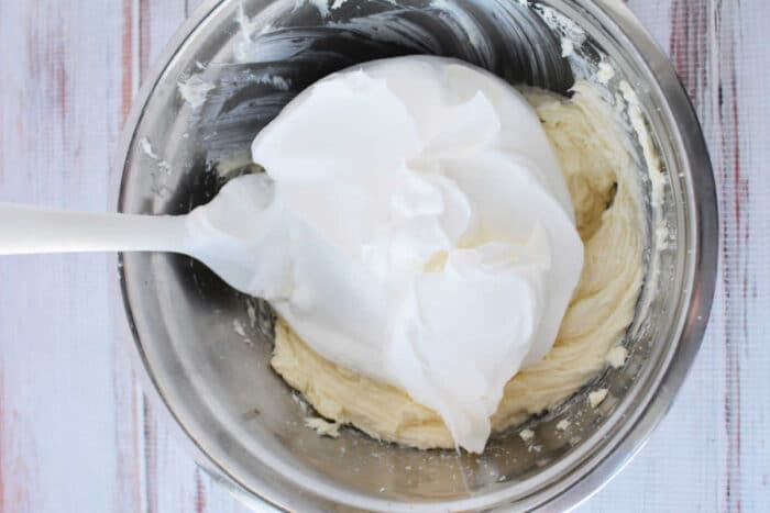 A white spatula folding fluffy egg whites into 3 ingredient cheesecake batter in a mixing bowl.