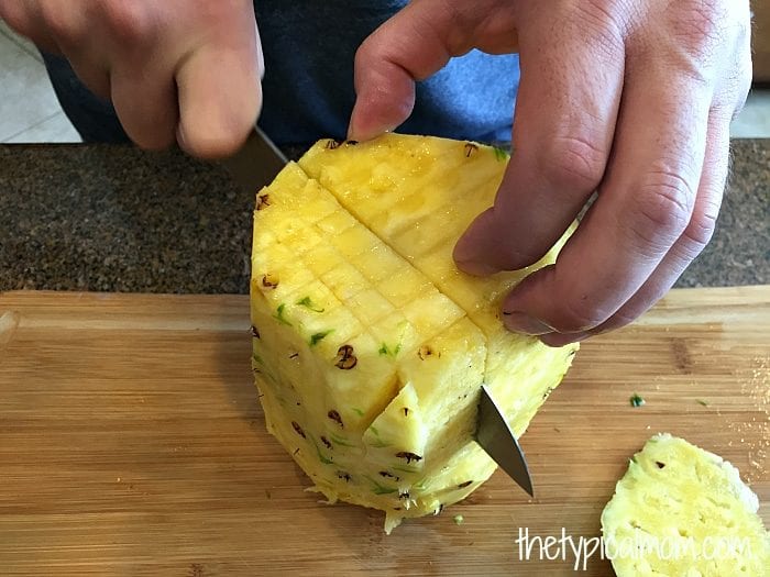 Hands using a knife to cut a pineapple into cubes on a wooden cutting board for a fruit salad with cool whip.