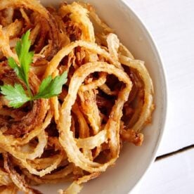 A white bowl of crispy fried onions, garnished with green parsley, sits on a white table.