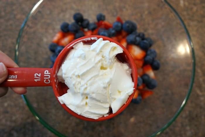 Easy fruit salad recipe that's super creamy and easy to make.. The perfect BBQ side dish or a light fruit dessert idea that everyone will love! A red measuring cup of cool whip held over a fruit salad of blueberries and sliced strawberries.