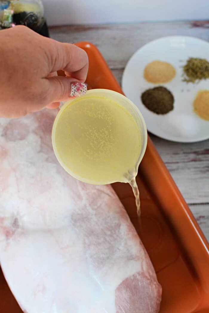 A hand pouring broth from a measuring cup onto a frozen pork loin in a baking pan.