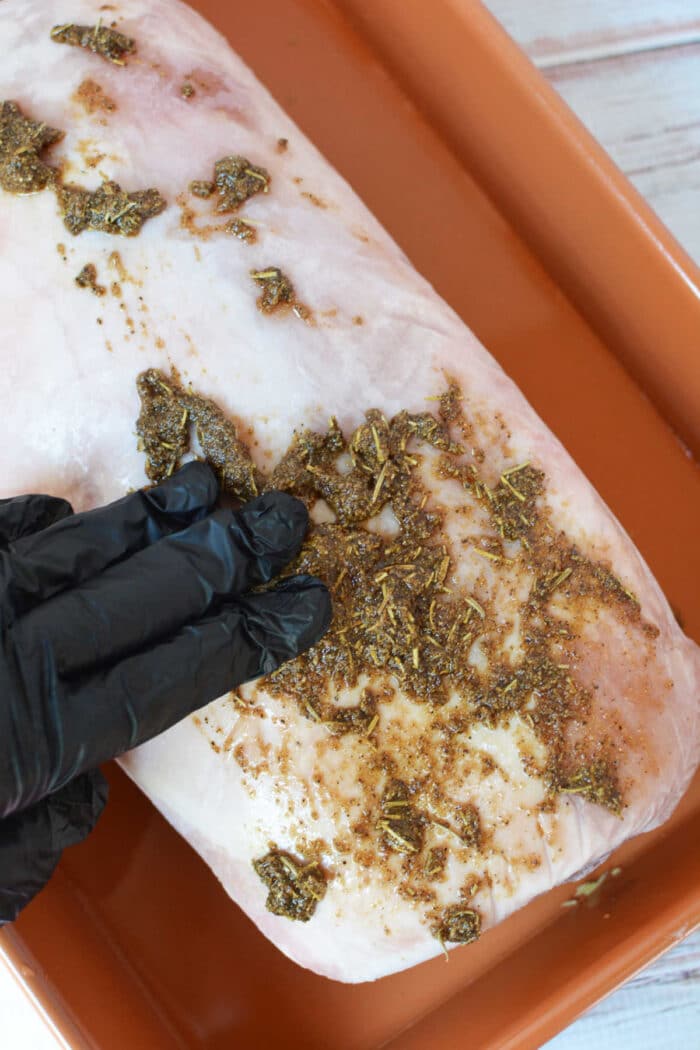 A gloved hand rubs dry herb seasoning onto a raw pork loin in a baking dish before it goes in the oven.